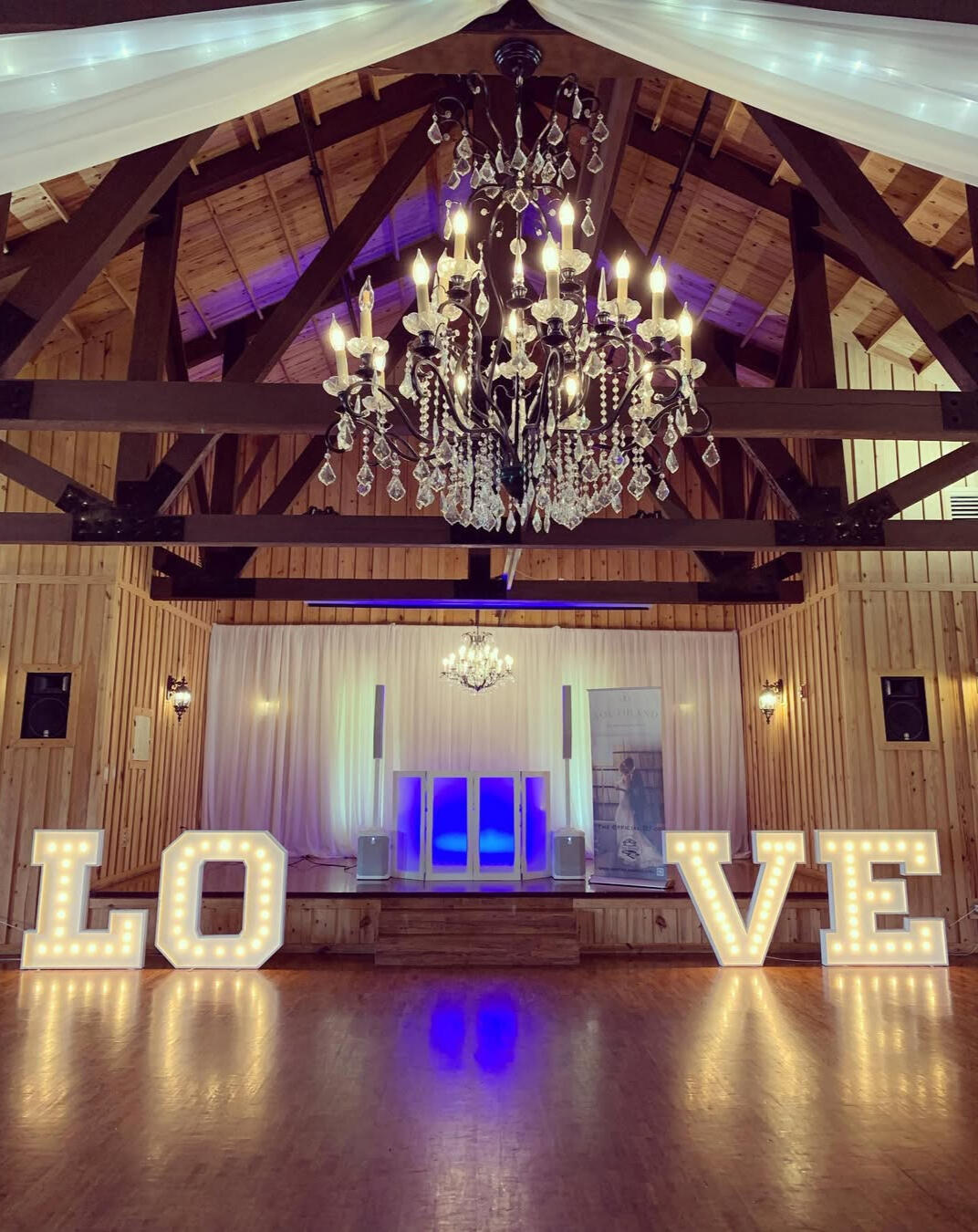 Wedding DJ Setup Large illuminated letters spelling "LOVE" stand on a wooden floor in a decorated hall with chandeliers, draped fabric, and wooden beams.
